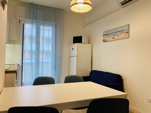 Une petite pièce moderne, avec une table blanche, deux chaises, un canapé bleu, un réfrigérateur, un tableau représentant une scène de plage et une fenêtre ornée de rideaux légers.