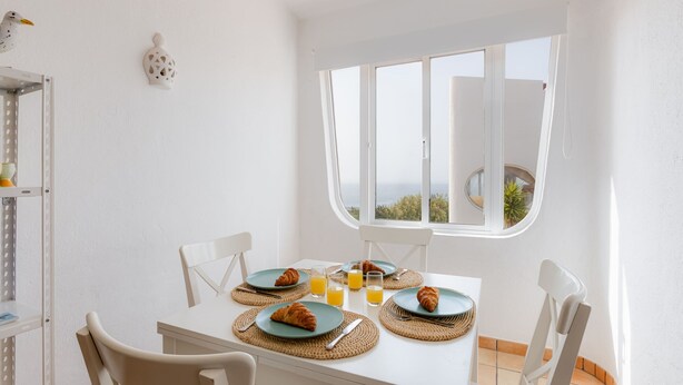 A dining area with a white table set for breakfast, including croissants and orange juice, with a view of the sea through a large window. - Solemar @ Vale de Centeanes (Carvoeiro)