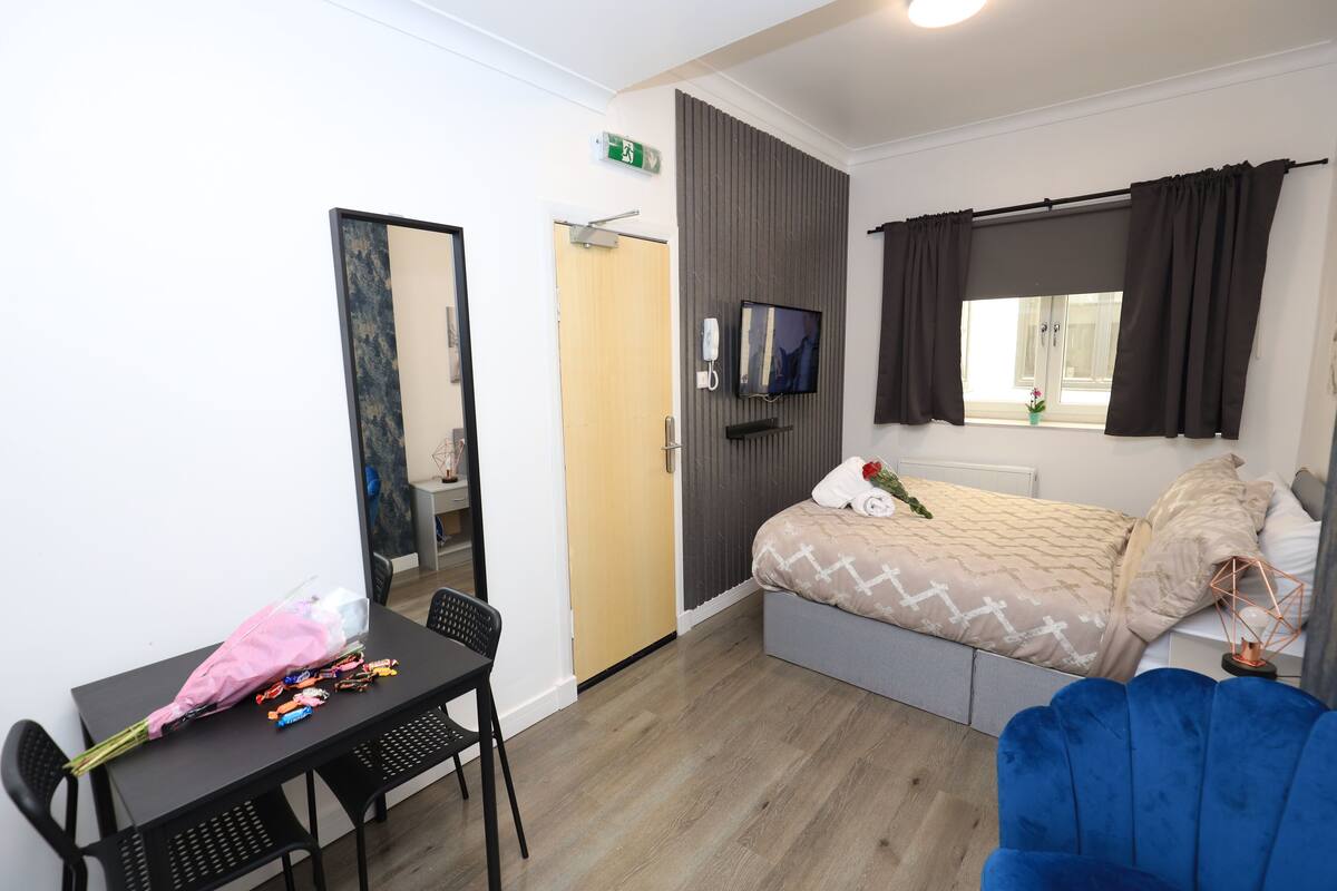 A modern hotel room with a bed, a TV, a desk with a chair, and a window with curtains.