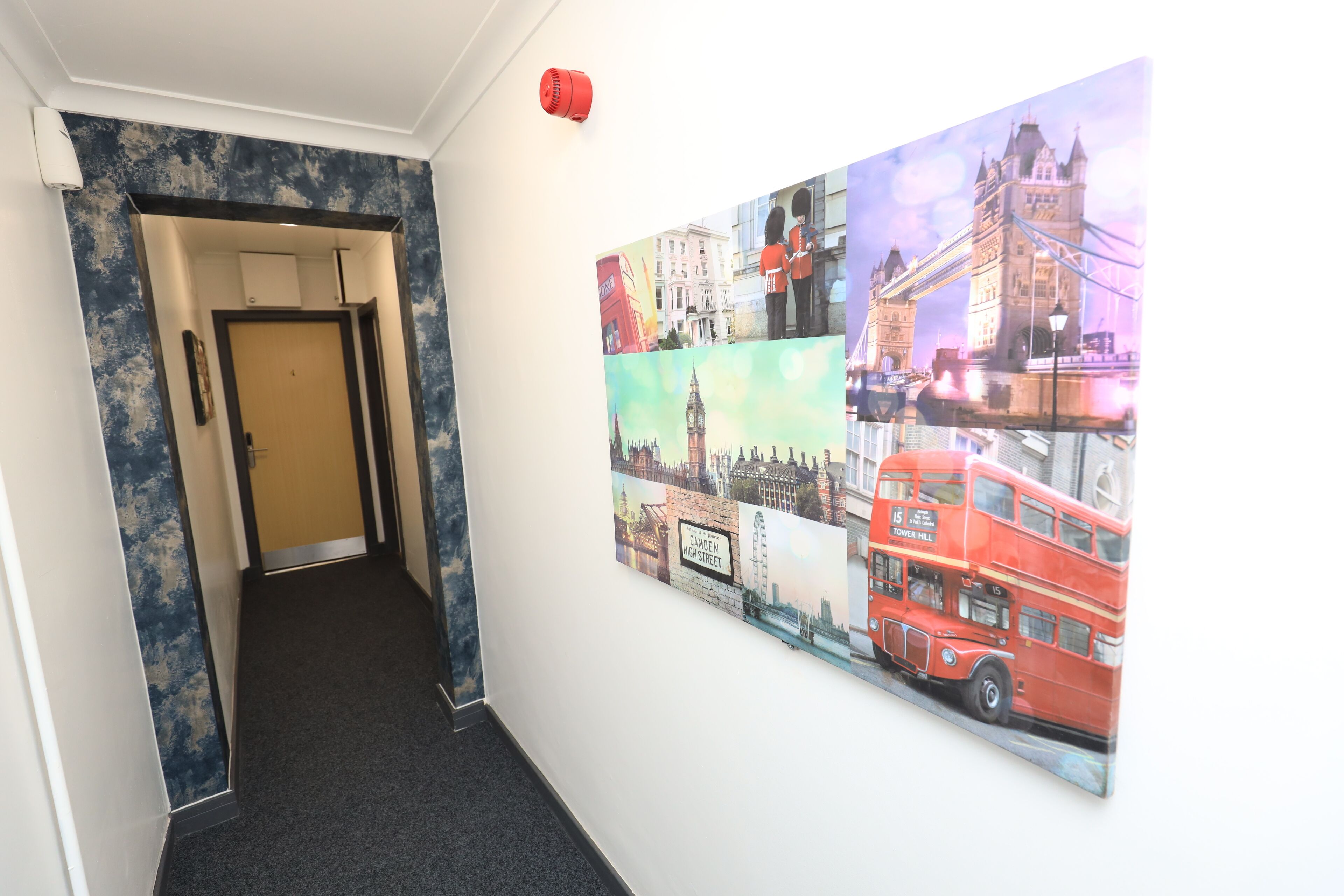 A hallway with a door at the end, a wall-mounted artwork depicting a red double-decker bus, and a red fire alarm on the wall.