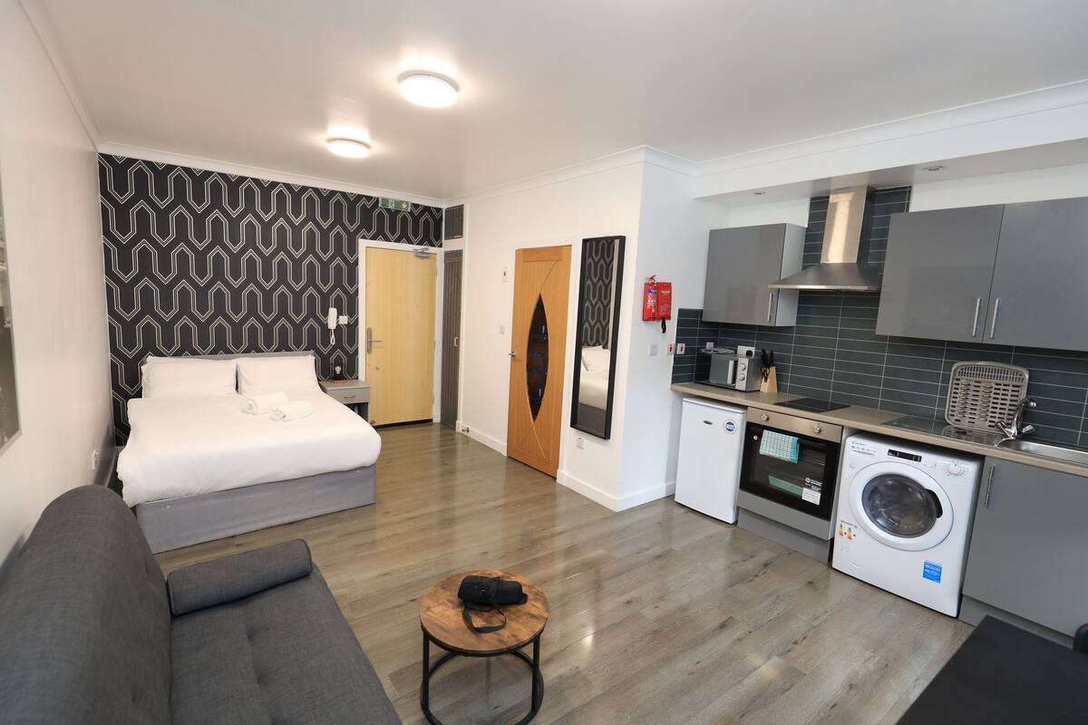 A compact hotel room with a bed, a sofa, a small table, a washing machine, and a kitchenette.