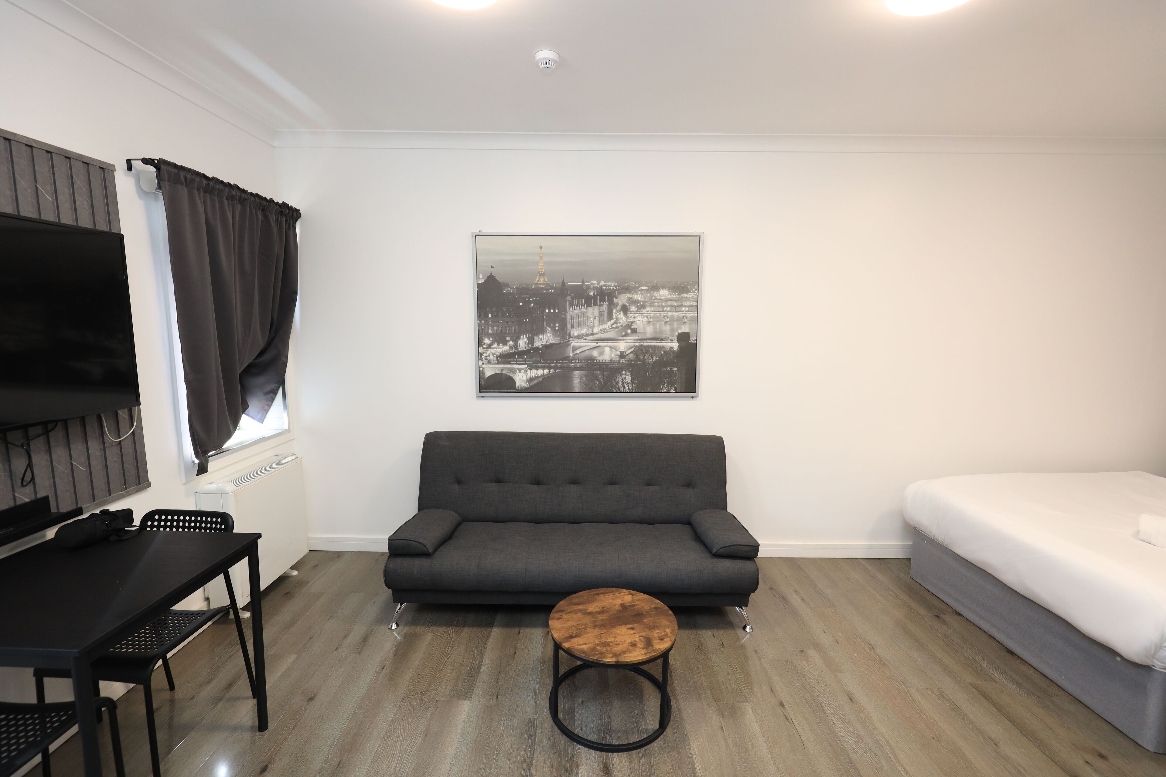 A modern room with a grey sofa, a small round wooden table, a bed, a TV, and a cityscape painting on the wall.