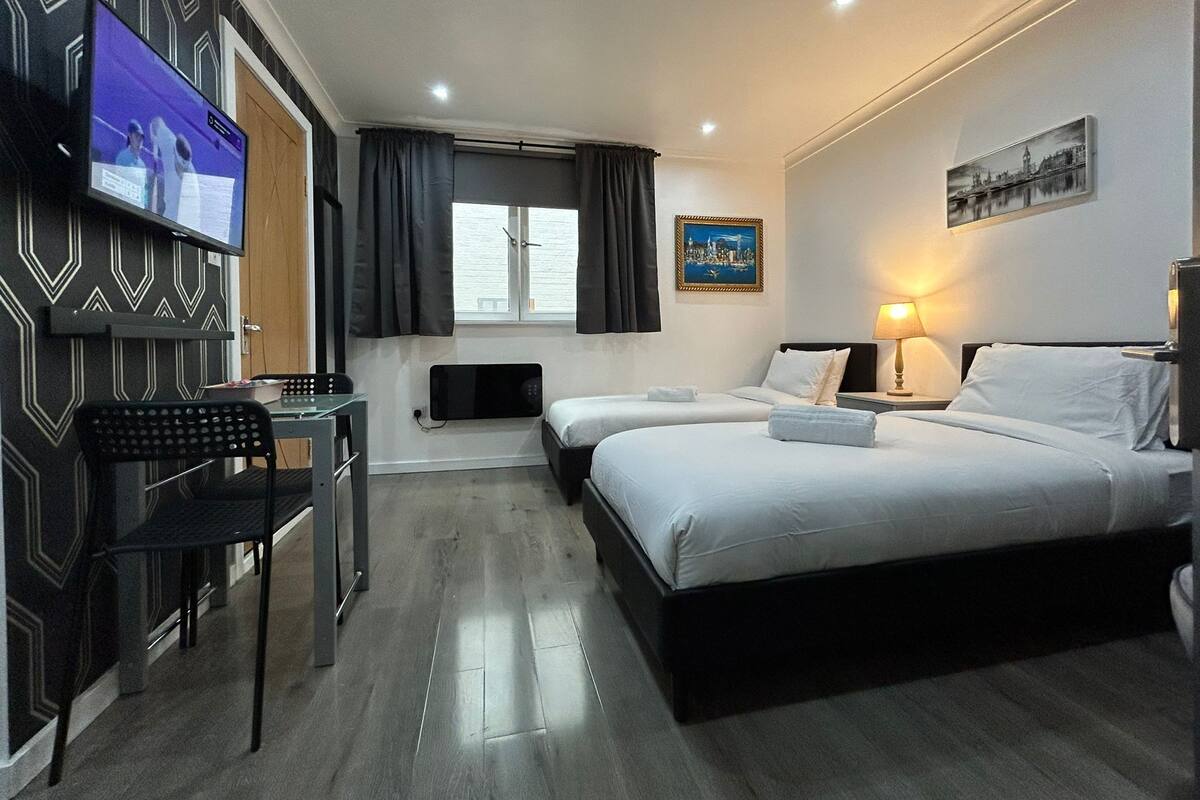A hotel room with two beds, a desk with a chair, a television, a window with curtains, and a painting on the wall.