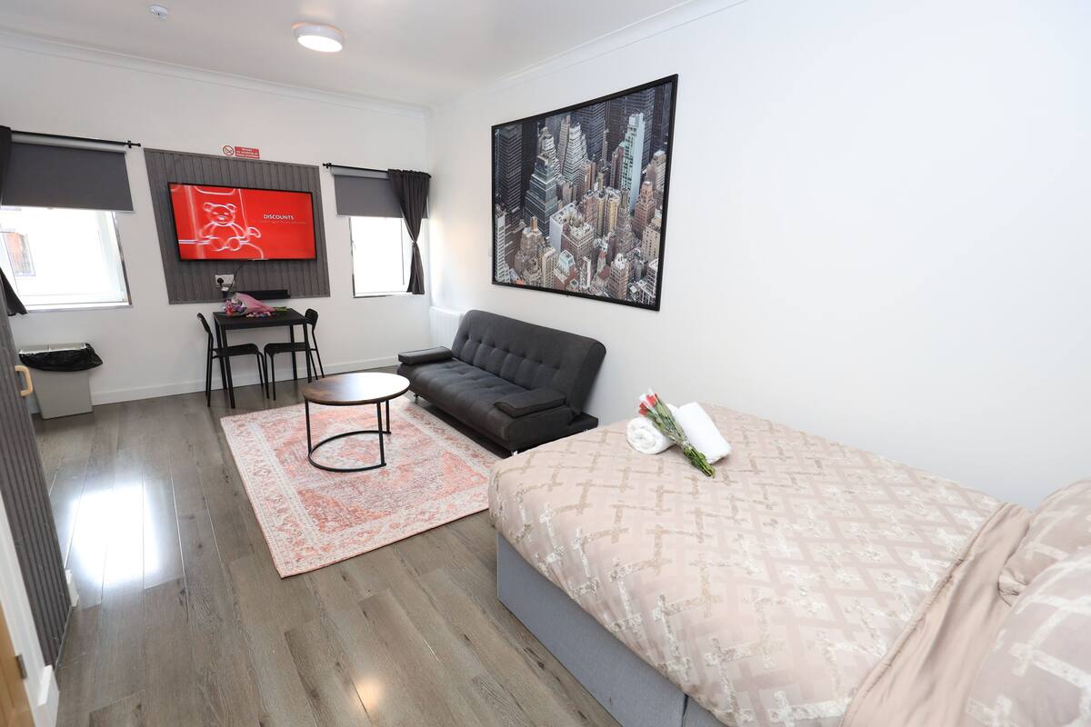 A modern hotel room with a bed, a sofa, a dining table with chairs, a flat-screen TV, and a cityscape painting.