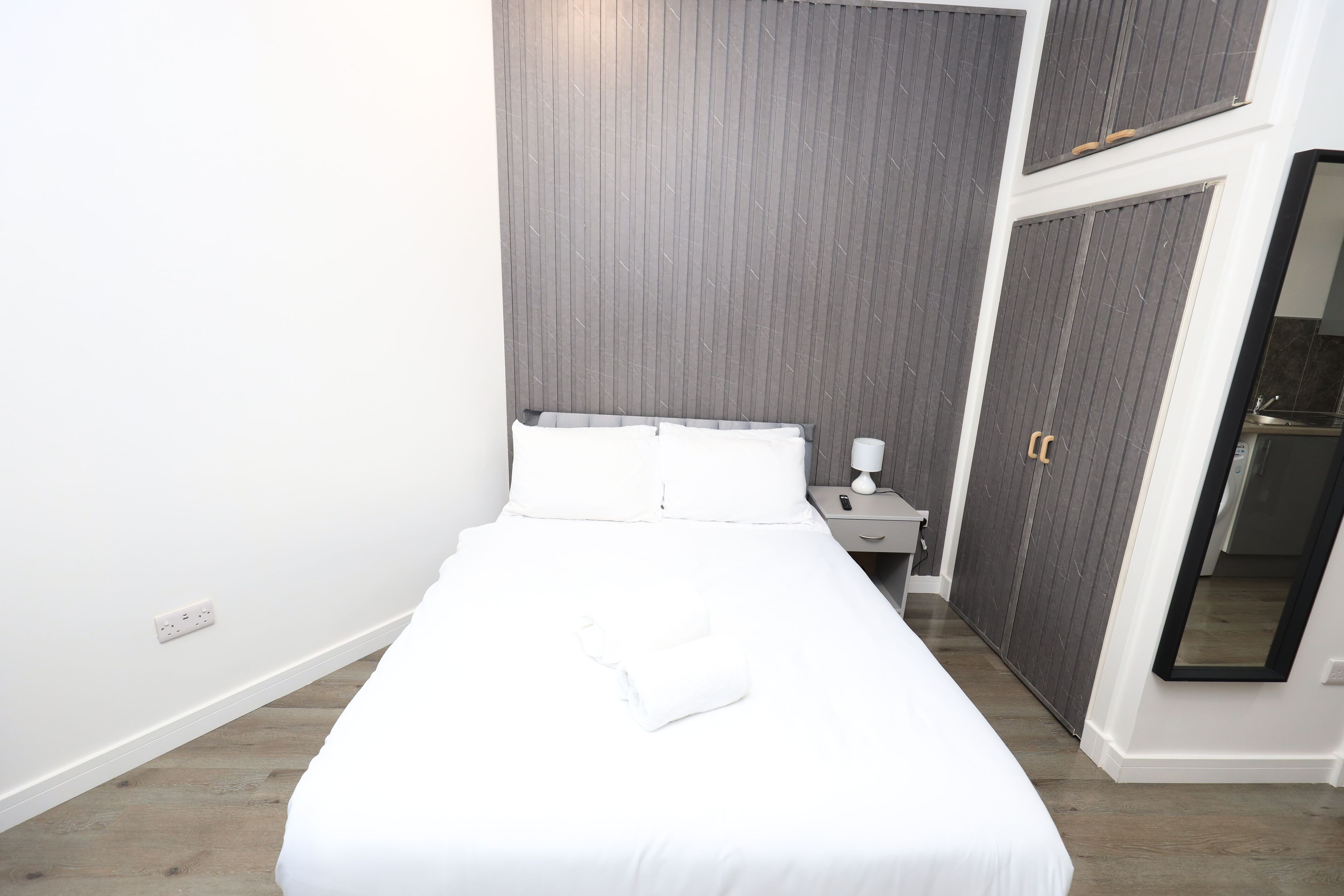 Un lit bien fait dans une chambre d’hôtel, avec un mur gris, une table de chevet avec une lampe et une armoire avec un miroir.