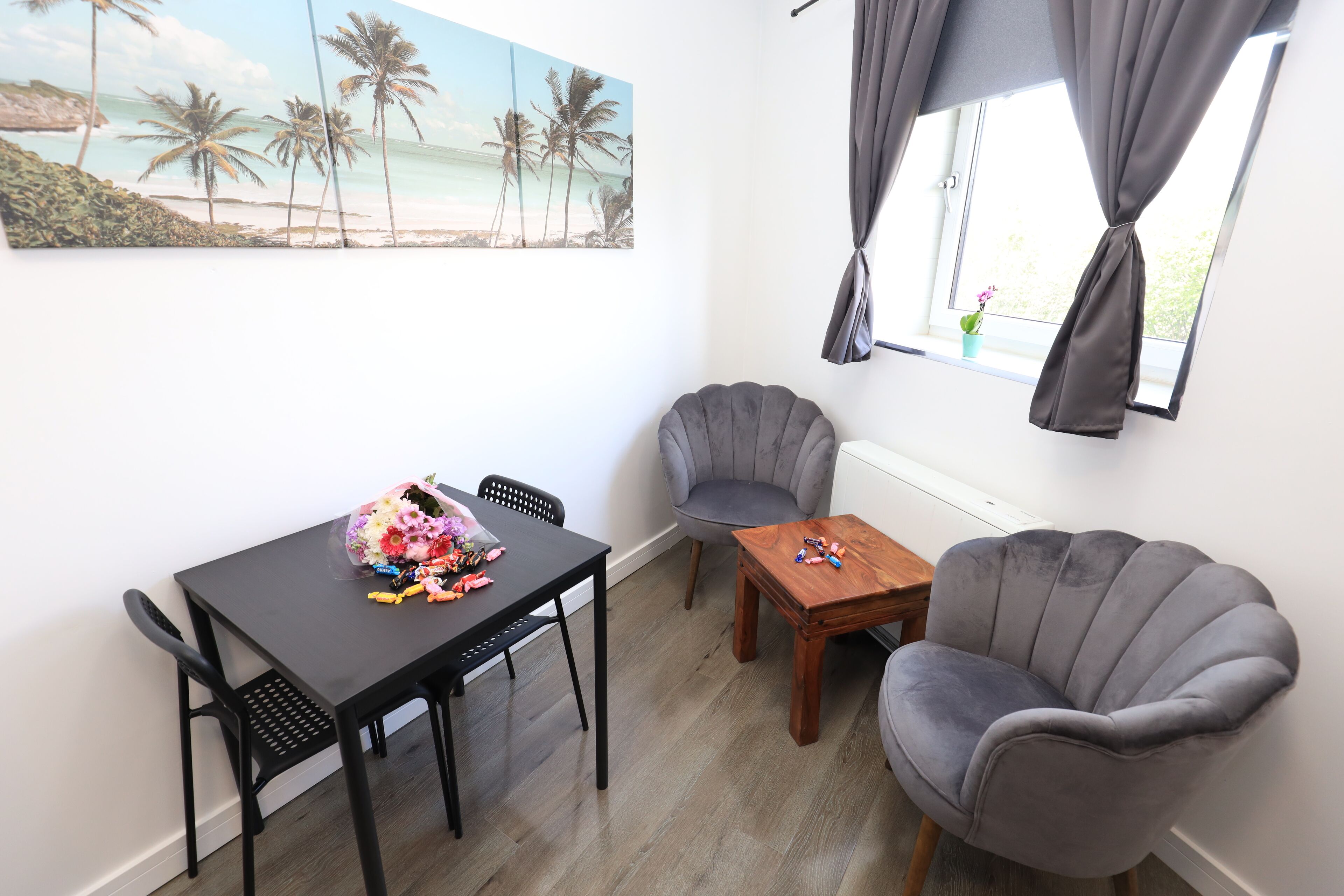 A room with a dining table, two chairs, a small wooden table, and two armchairs. A painting of a beach scene is on the wall.