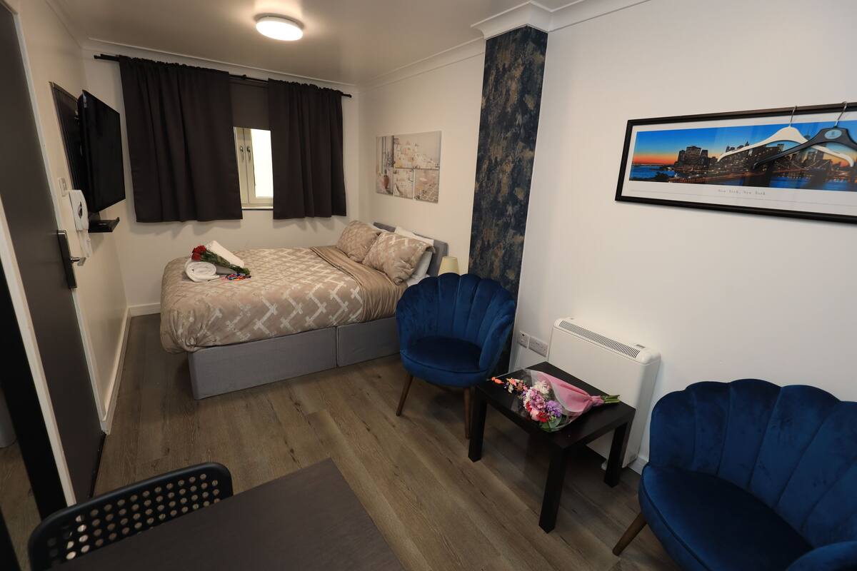 A bedroom with a bed, two blue chairs, a small table with flowers, a framed picture of a cityscape, and a window with curtains.