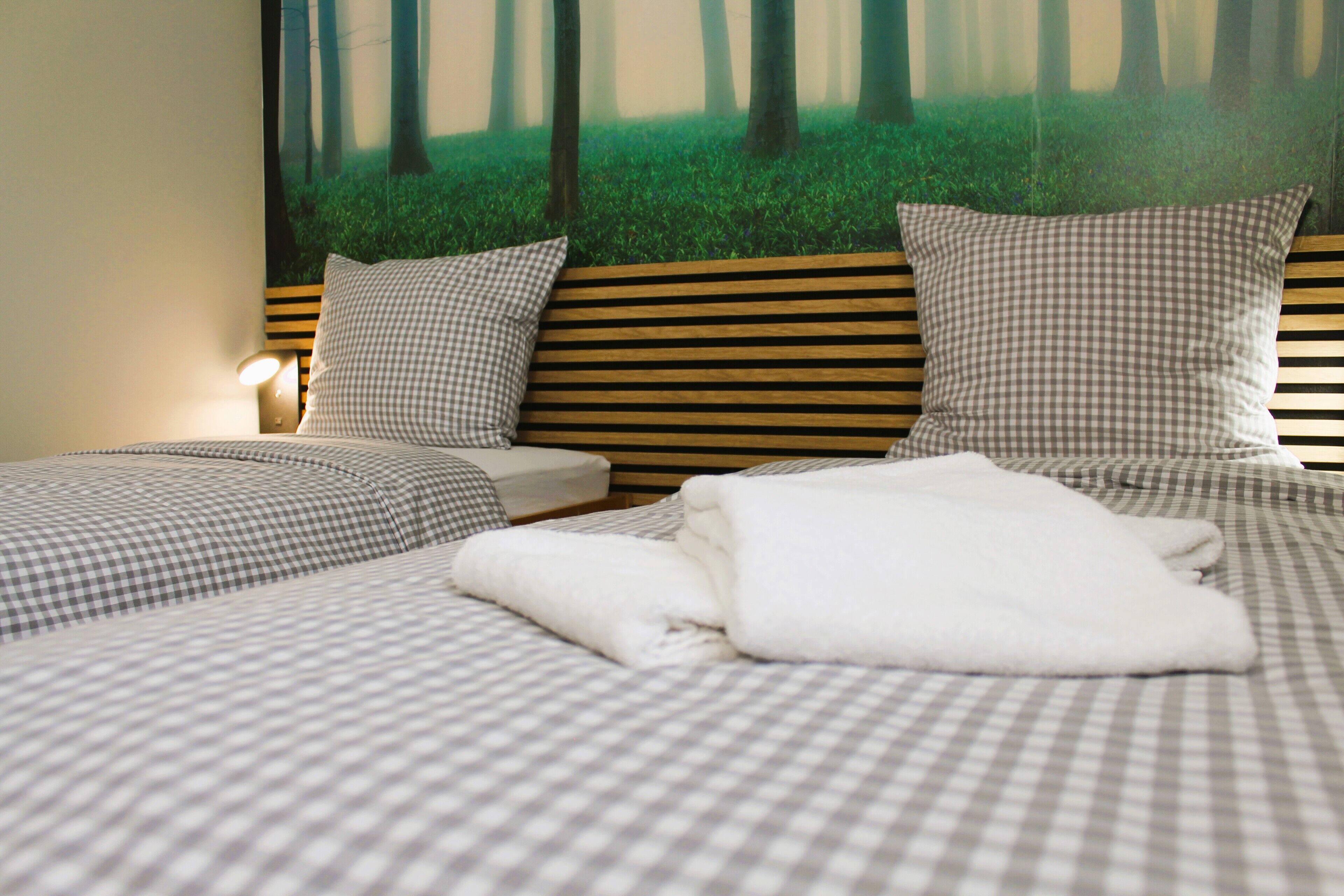 A neatly made bed with checkered bedding and pillows, a bedside table with a lamp, and a wall mural of a forest scene.