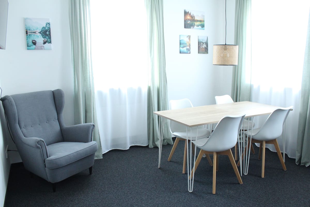 A modern room with a grey armchair, a wooden table with white chairs, and a hanging light fixture.