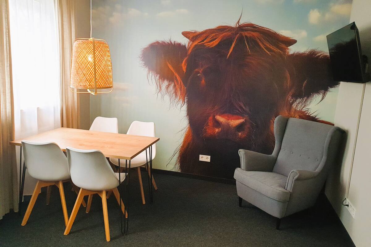 A conference room with a large mural of a cow, a wooden table with chairs, a grey armchair, and a hanging lamp.
