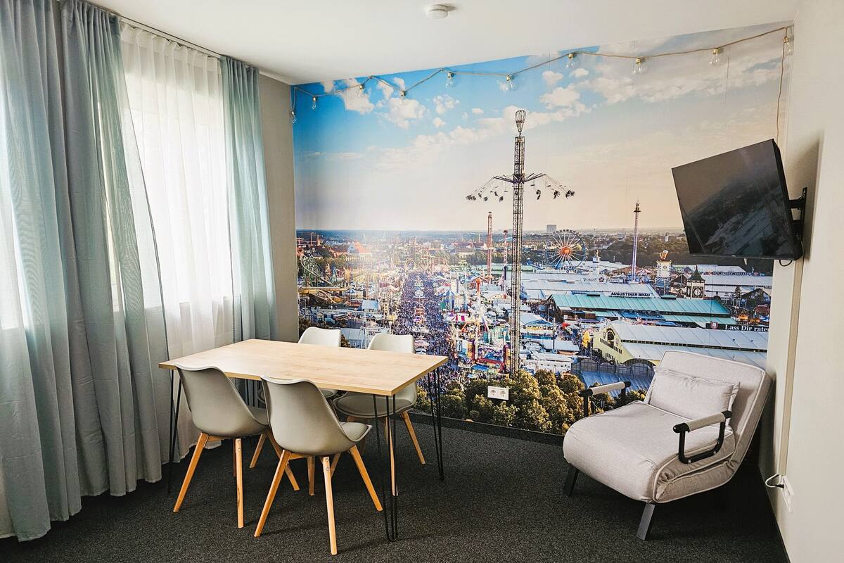 A hotel room with a cityscape mural, a dining table with chairs, a sofa, and a TV mounted on the wall.