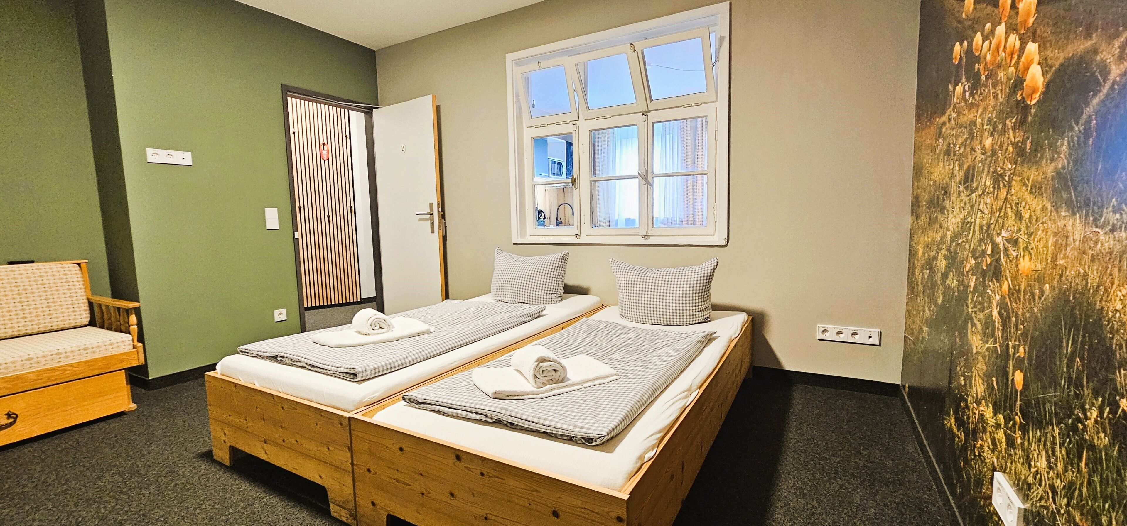 Una habitación con dos camas individuales, un banco de madera, una pared verde, una ventana y una obra de arte colgada en la pared.
