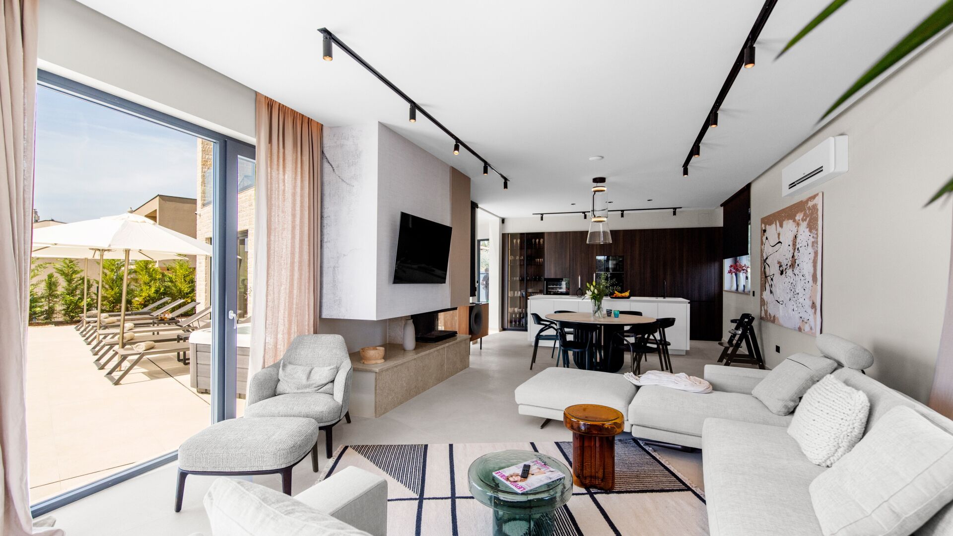 Un salon moderne comprenant un canapé, des fauteuils, une table basse et un téléviseur. Il y a également un coin repas avec une table et des chaises. La pièce est dotée de grandes fenêtres et offre une vue sur l’extérieur.