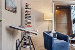Een kamer met een tekentafel, een blauwe fauteuil, een lamp en een boekenplank tegen de muur.