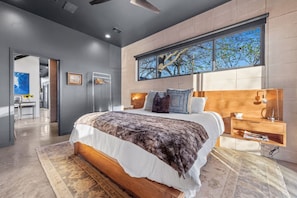 Une chambre moderne avec un grand lit, une tête de lit en bois, une table de chevet et une fenêtre donnant sur un arbre.