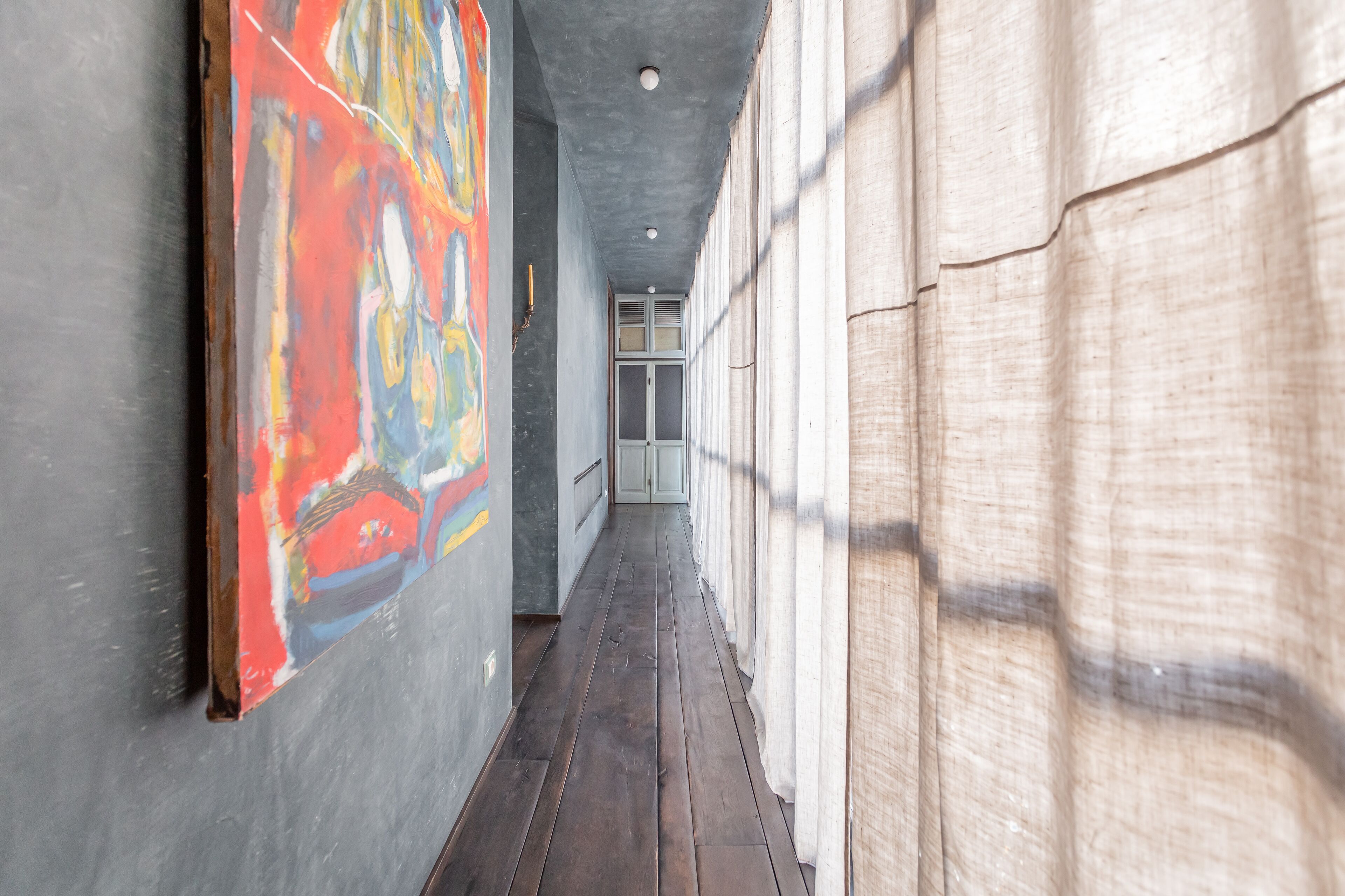 A narrow hallway with a wooden floor, a colorful abstract painting on the left, and a plain wall on the right.