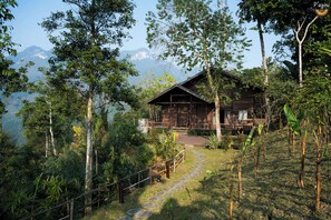 A wooden cabin with a stone pathway leading to it, surrounded by lush greenery and mountains in the background.
