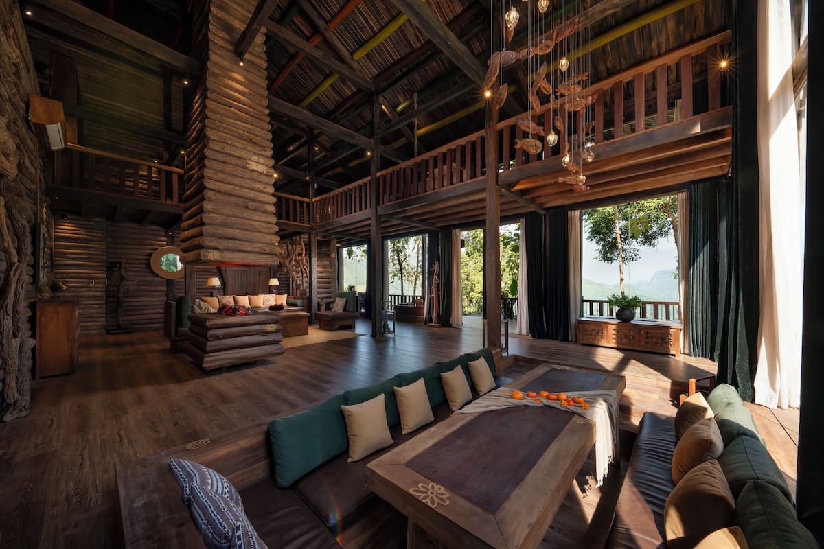 A spacious log cabin interior with a large wooden dining table, multiple seating areas, and a balcony with a view of the outdoors.
