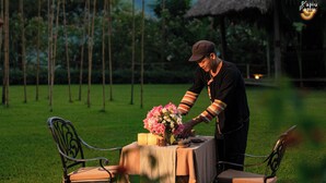 A person arranging a table with a candle and flowers in an outdoor setting with chairs and a thatched structure in the background.