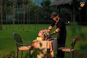 A person arranging a table with a candle and flowers in an outdoor setting with chairs and a thatched structure in the background.