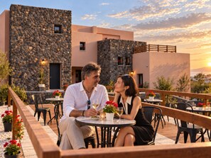A couple dining at an outdoor table with a stone wall backdrop, surrounded by potted plants and a wooden deck. -  Golden Moments Luxury Stone Villas. (Santorini)