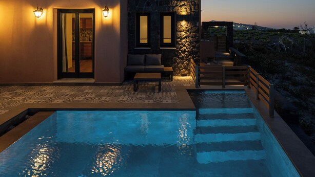 A modern building with a swimming pool, stone wall, and outdoor seating area. -  Golden Moments Luxury Stone Villas. (Santorini)