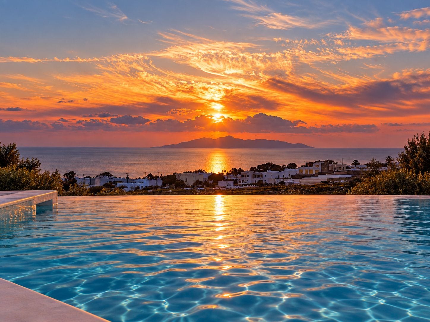 A swimming pool with clear water reflecting the sunset, a coastal town in the background, and a mountain range across the water.