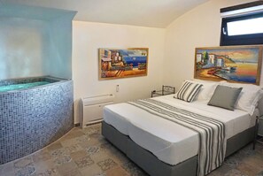 A bedroom with a bed, a small window, a wall-mounted heater, and a tiled bathtub. -  Golden Moments Luxury Stone Villas. (Santorini)