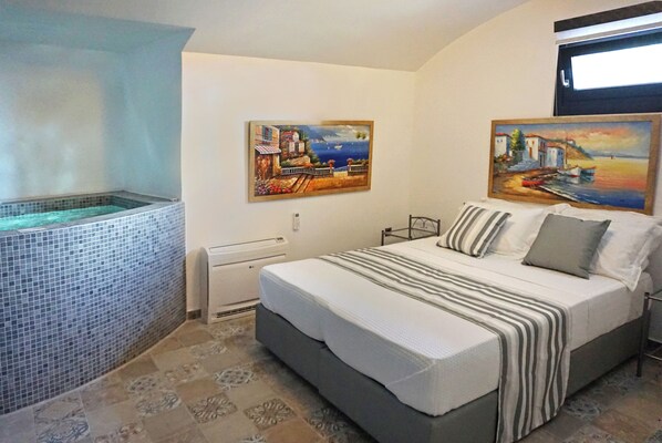 A bedroom with a bed, a small window, a wall-mounted heater, and a tiled bathtub. -  Golden Moments Luxury Stone Villas. (Santorini)