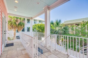 A covered balcony with white railings, a stone floor, and a view of a building with a green roof and palm trees. - Game Room and Private Heated Pool + 5BR Sleeps 12 (Tybee Island)