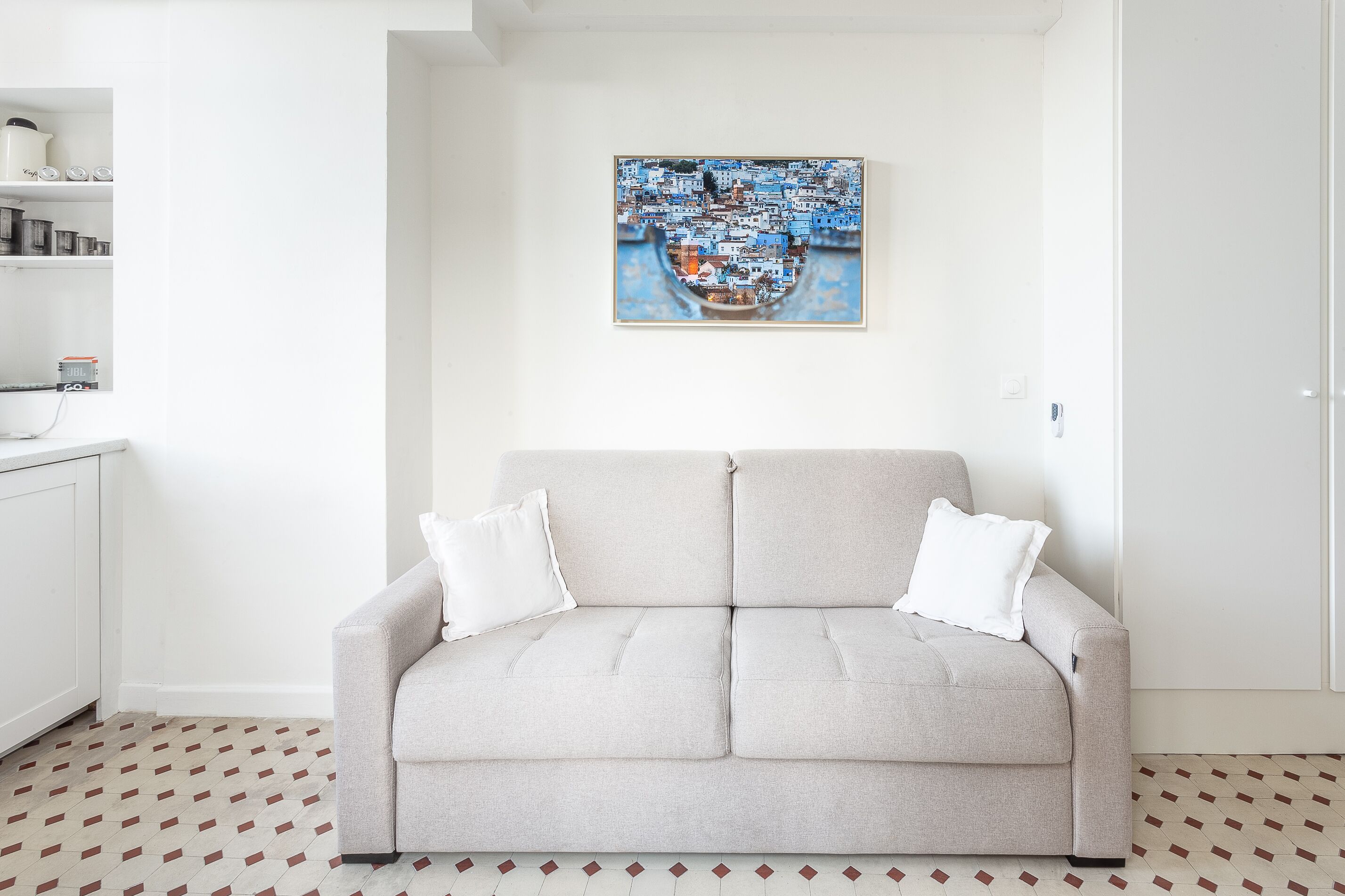 Un soggiorno con un divano grigio chiaro, due cuscini bianchi, un quadro appeso al muro e un pavimento a motivi.