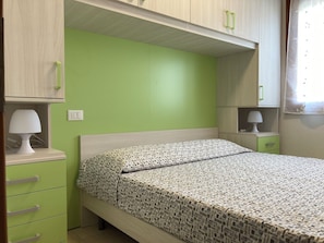A bedroom with a bed, a nightstand, a lamp, a drawer unit, and a window with curtains. - Lovely Flat in the Center of Rosolina Mare (Rosolina)