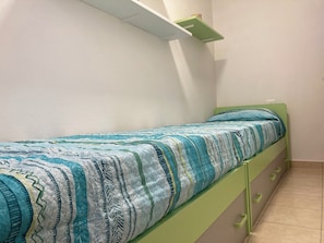 Ein Zimmer mit einem Einzelbett, einem grünen Bettgestell, einer gemusterten Bettdecke und einer weißen Wand.