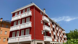 Un immeuble résidentiel à plusieurs étages, doté de balcons et d’une façade rouge, avec un ciel bleu clair en arrière-plan.