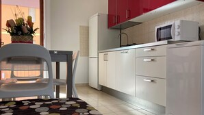 Une cuisine compacte avec des meubles blancs, un four à micro-ondes et un réfrigérateur. On y trouve une table à manger avec une chaise et un tapis décoratif.