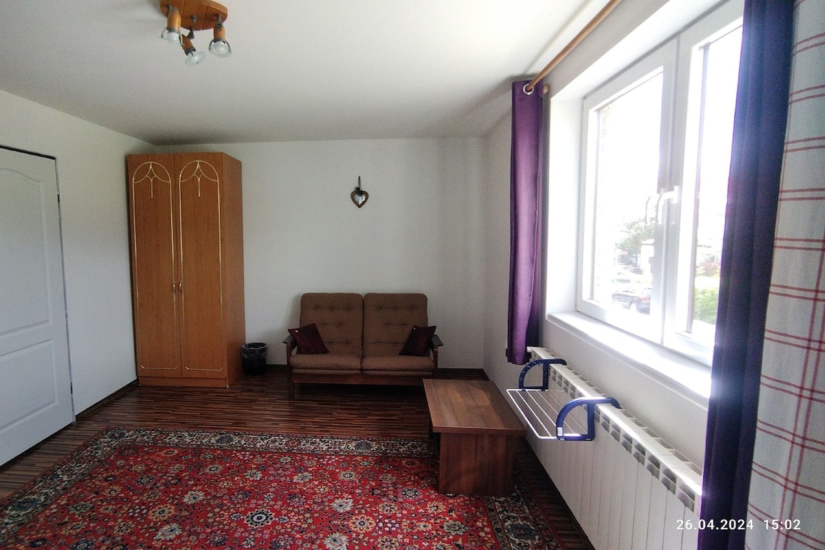 A room with a wooden wardrobe, a sofa, a patterned rug, a small table, and a window with curtains.