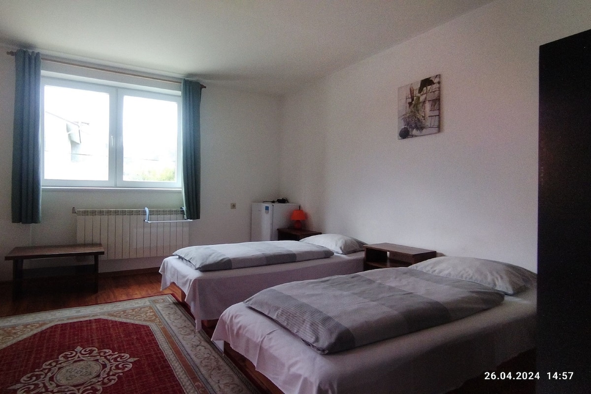A room with two single beds, a small table, a bench, a radiator, and a window with curtains.