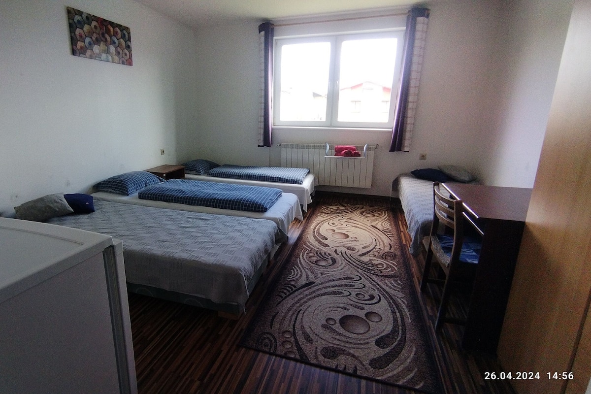 A small, clean room with two single beds, a desk, a lamp, and a window with curtains.