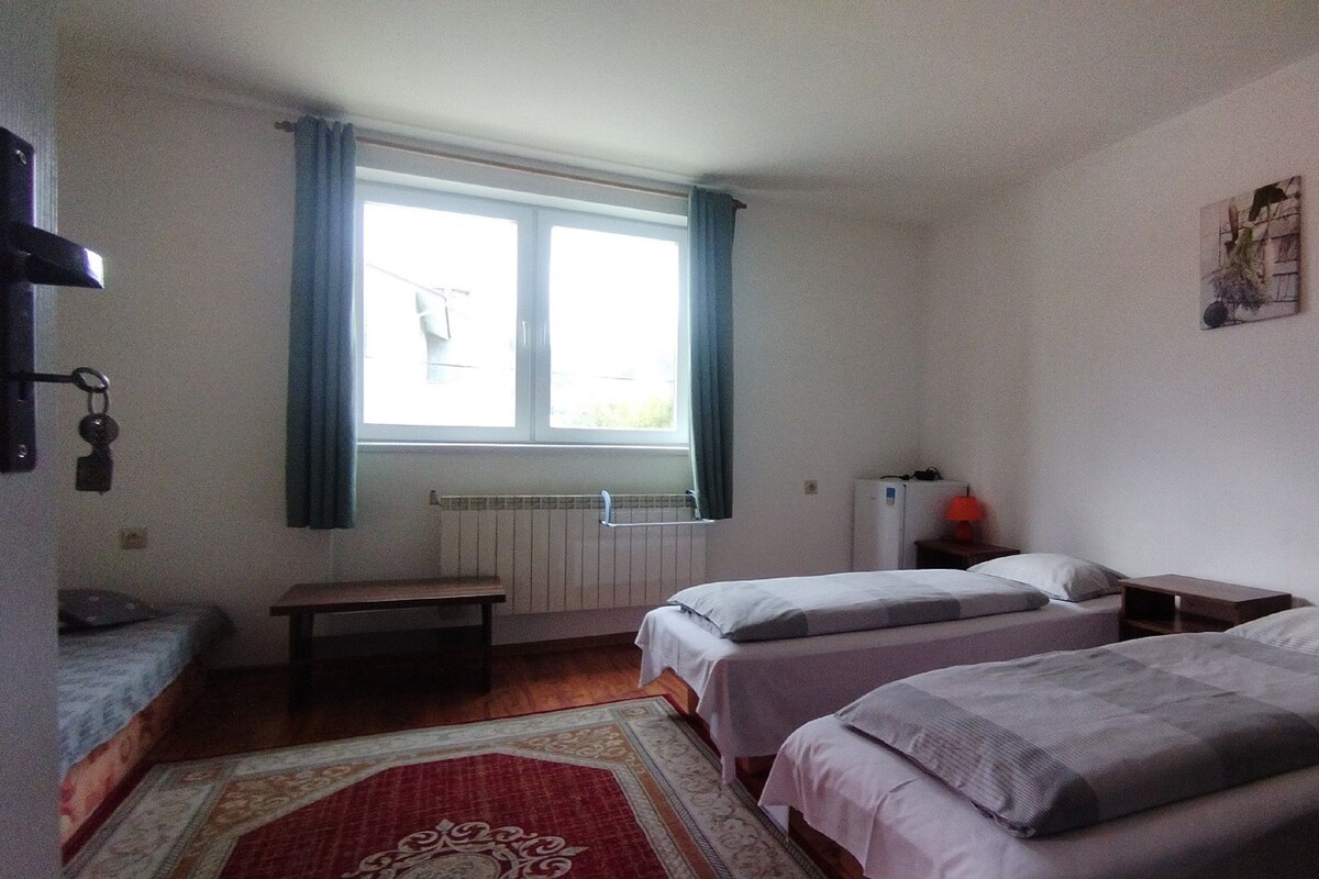 A room with two single beds, a small table, a bench, a radiator, and a window with curtains.