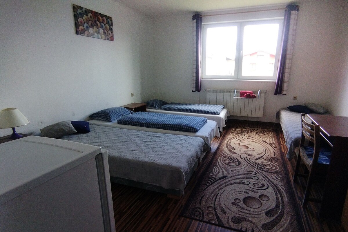 A small, clean room with two single beds, a desk, a lamp, and a window with curtains.