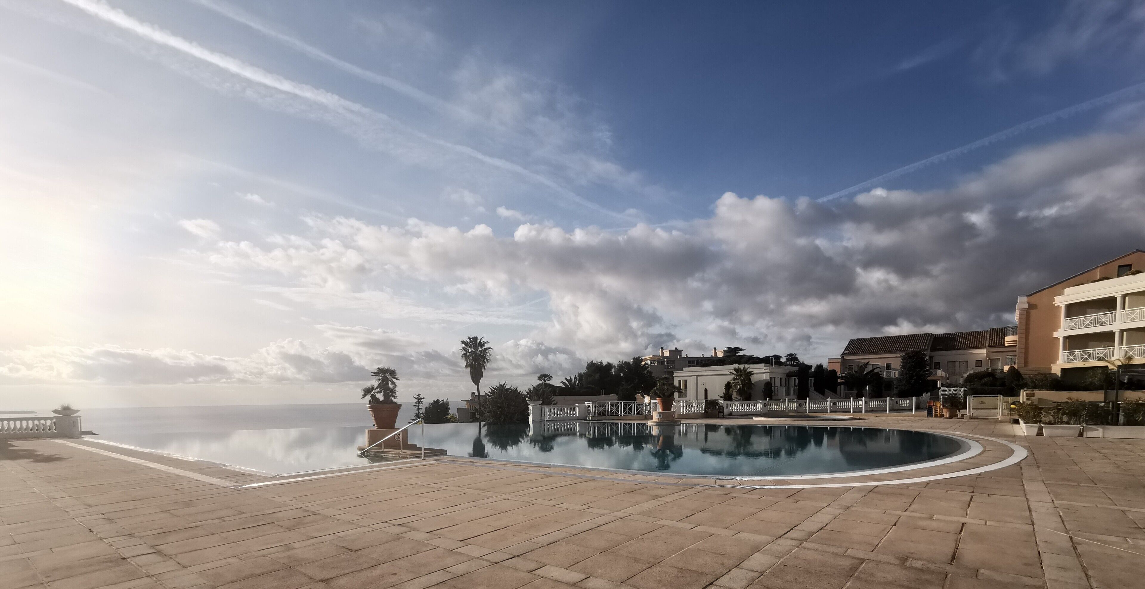 A waterfront pool area with a clear view of the sea, surrounded by buildings and potted plants.