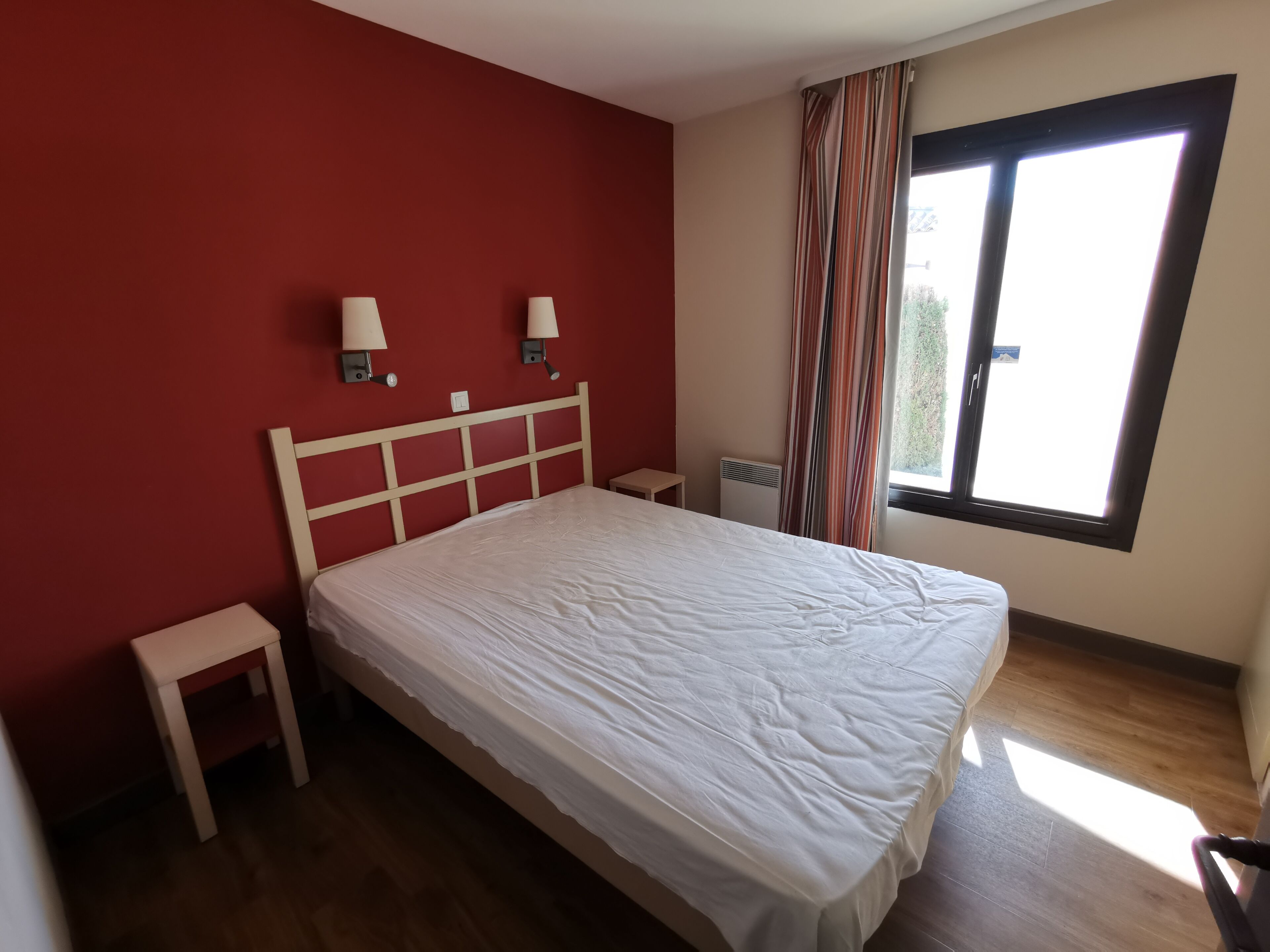 A bedroom with a bed, two bedside tables, a window with curtains, and a radiator.