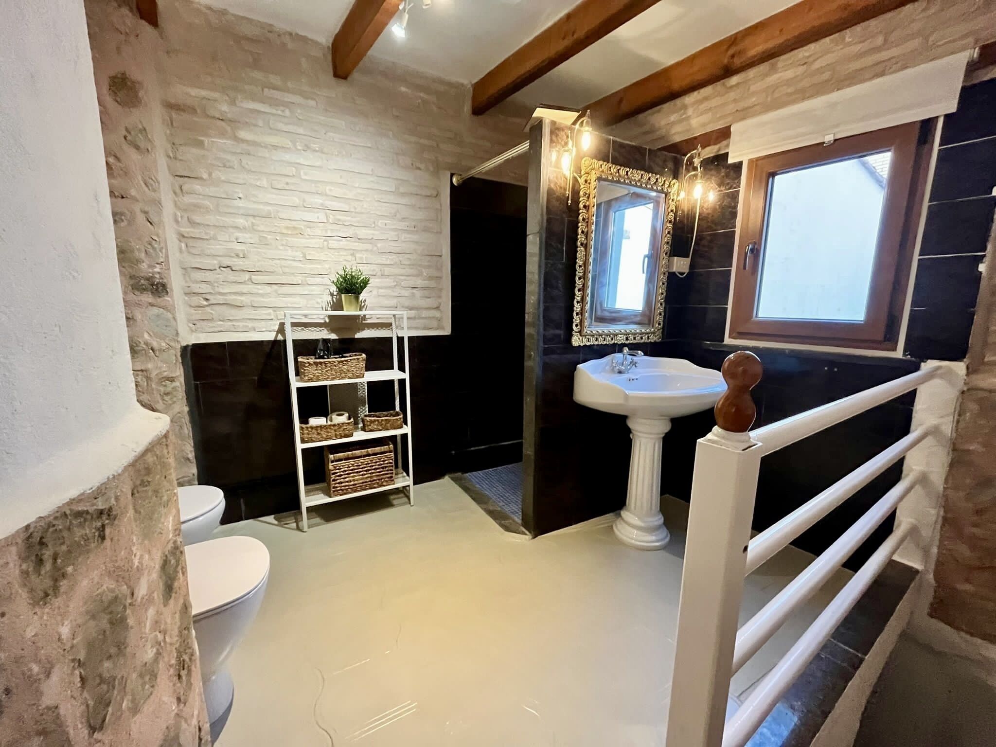 Un baño con pared de ladrillo blanco, un lavamanos pedestal blanco, un taburete de madera, un estante blanco con cestas, y una ventana con vistas.