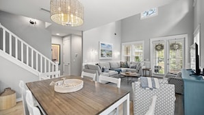 A spacious living room with a wooden dining table, white chairs, a grey sofa, and a blue cabinet.