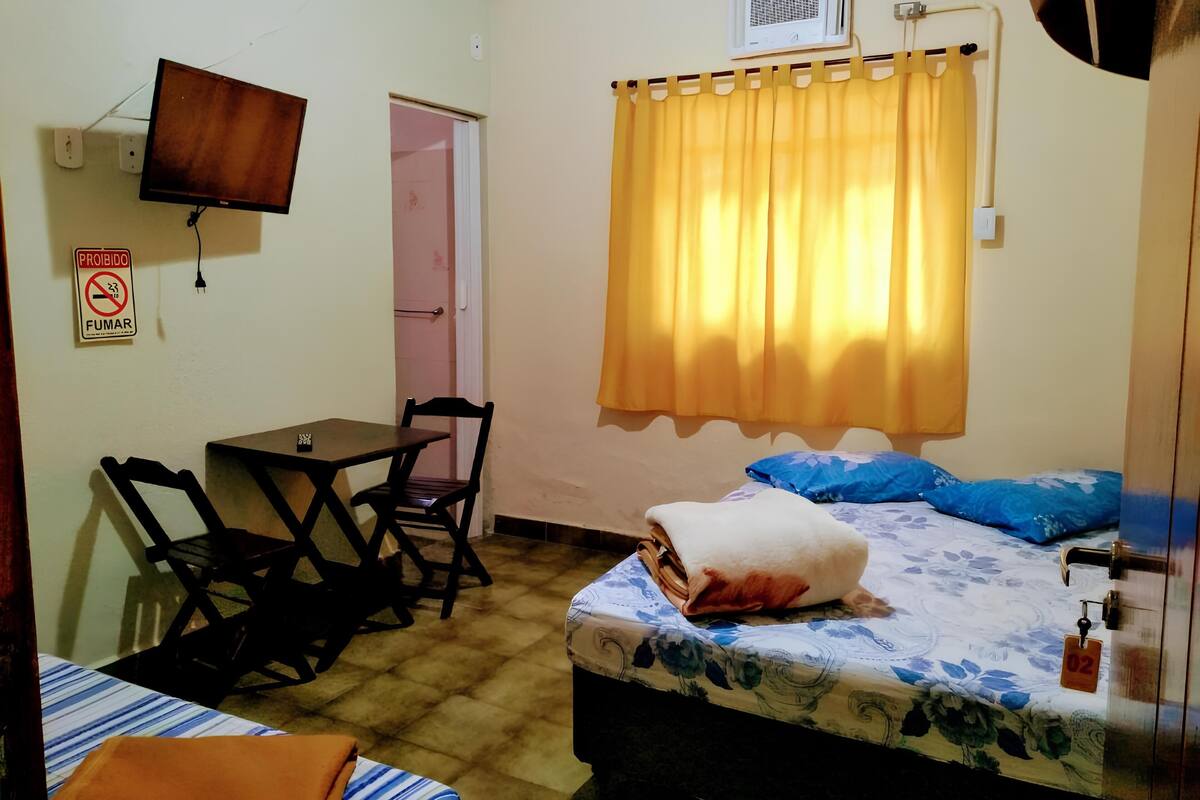 A small room with a bed, a desk, and a chair. There is a television mounted on the wall, a window with curtains, and a door.