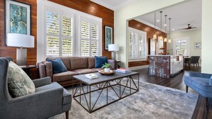 A spacious living room with a brown leather sofa, a grey armchair, and a coffee table. There are large windows with white shutters, a kitchen area with a bar, and a dining table.