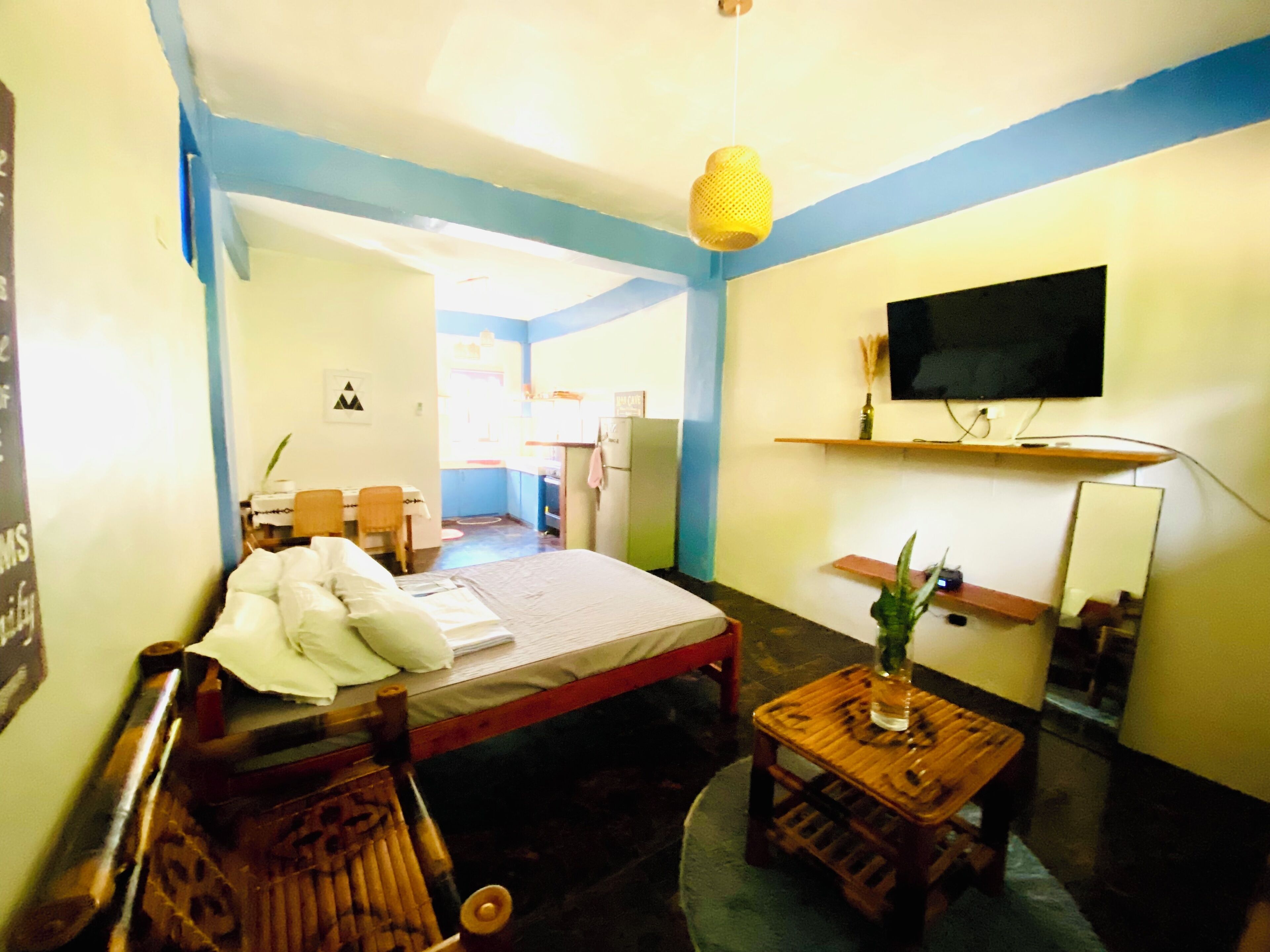 Una habitación con cama, una mesa de madera con una planta, un televisor en un estante y una pequeña cocina al fondo.