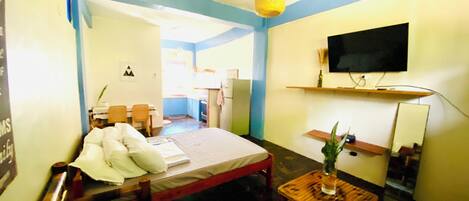 A room with a bed, a wooden table with a plant, a television on a shelf, and a small kitchen area in the background.