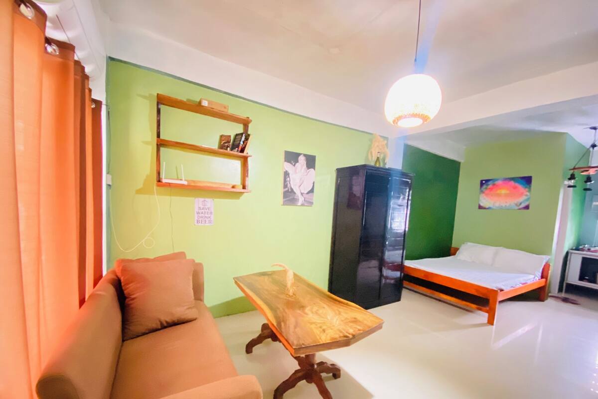 A room with a sofa, a wooden table, a bed, a black wardrobe, and a green wall with a 'Save Water Drink Beer' sign.