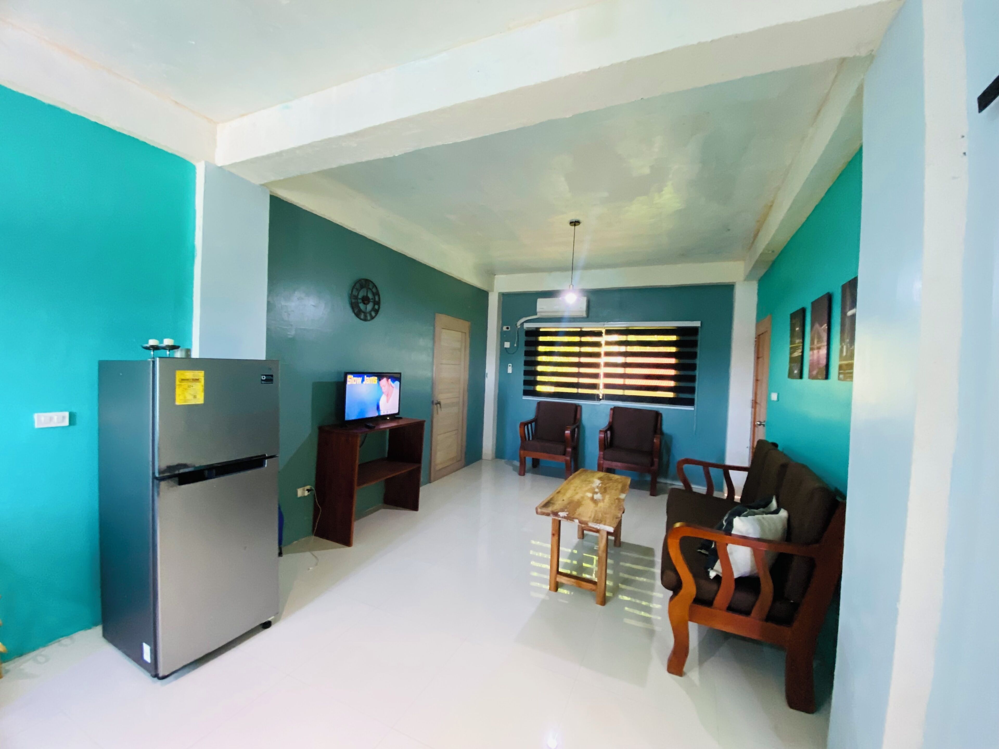 Un espacio habitable compacto con paredes verde azulado, un refrigerador, un televisor, una mesa de madera y dos sofás.